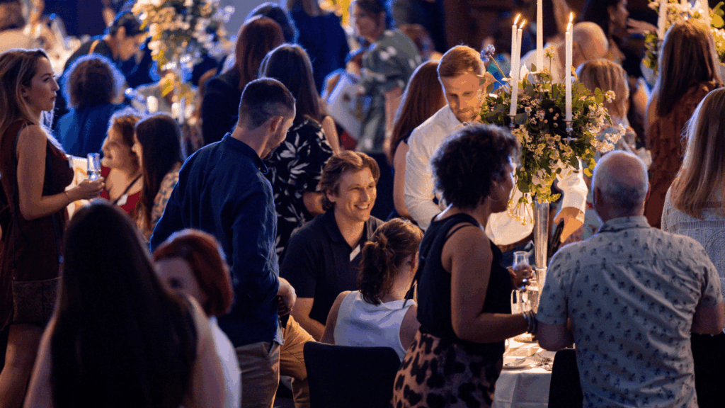 A lively private luxury event with people socializing around decorated tables, featuring tall floral centerpieces and lit candles. Guests are dressed in semi-formal attire, engaged in conversation and holding drinks.