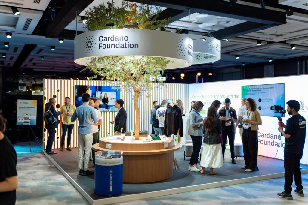 People interact and network at a Cardano Foundation exhibit booth, enhanced by Event Marketing Services, featuring a round table, informational screens, and a tree centerpiece within a modern, well-lit conference or expo setting.