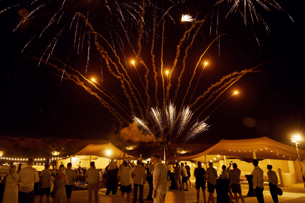 A crowd of people at an outdoor incentive travel event watch a colourful fireworks display in the night sky, with fairy lights and marquees illuminating the scene below.