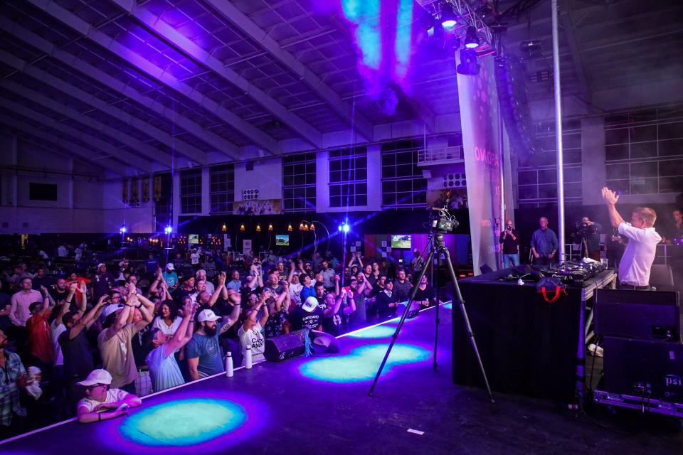 A DJ performs on stage inside a large venue, facing a cheering crowd under purple and blue lights. Fans raise their hands and take photos, while creative event services ensure the atmosphere is unforgettable. Ceiling and windows are visible in the background.