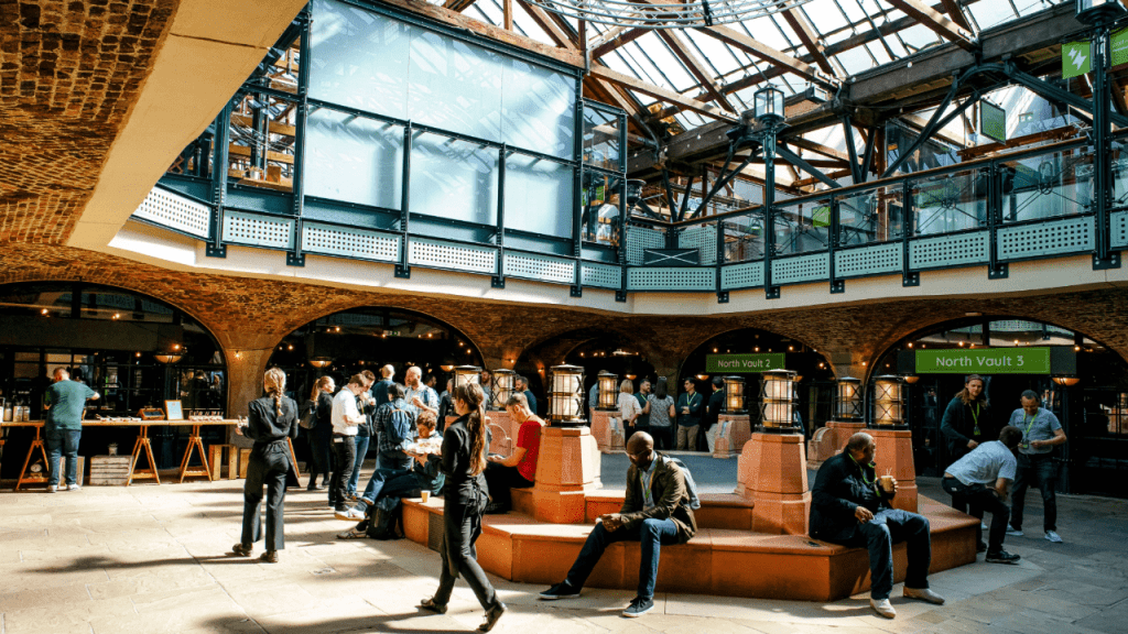 People gather and converse in a spacious, sunlit atrium with brick arches, modern glass walls, and signs reading “North Vault 2” and “North Vault 3.” This unique venue is a hidden gem often discovered by savvy event space finders.