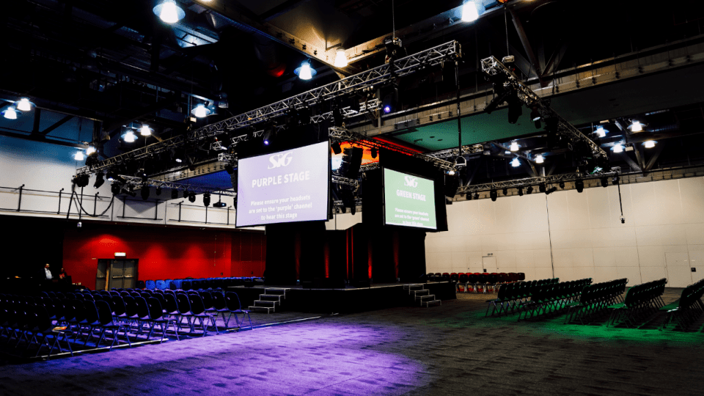 A large conference room with rows of empty chairs facing a central stage, above which hang large screens displaying “SVG Purple Stage” and event information. This vibrant setting is perfect for those using an event space finder to plan memorable gatherings.