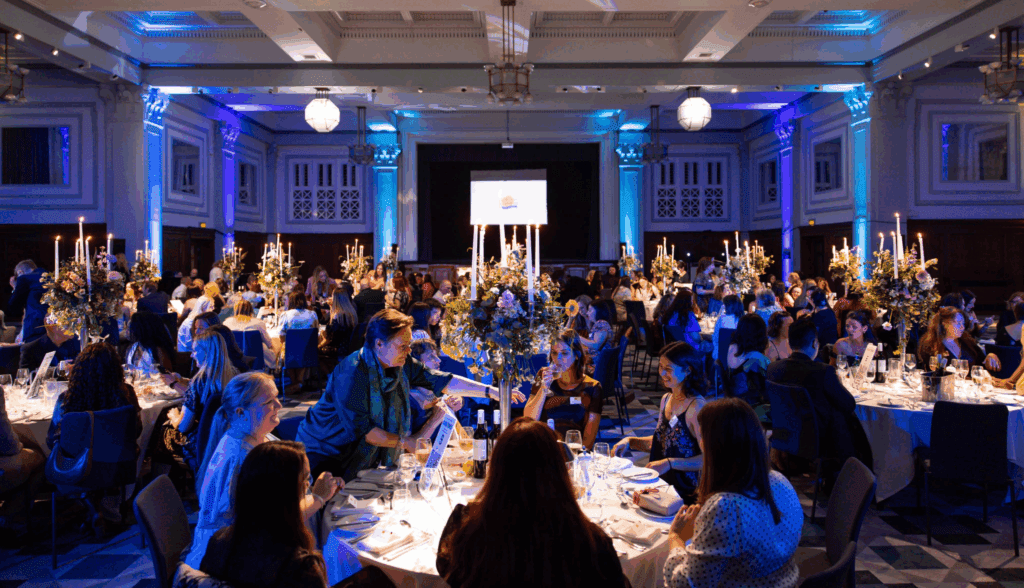 A private luxury event unfolds in a grand hall, where elegant lighting and lavish décor set the scene. Guests in formal attire dine and converse at round tables adorned with tall floral displays and candles, as a screen showcases the logo at the front.
