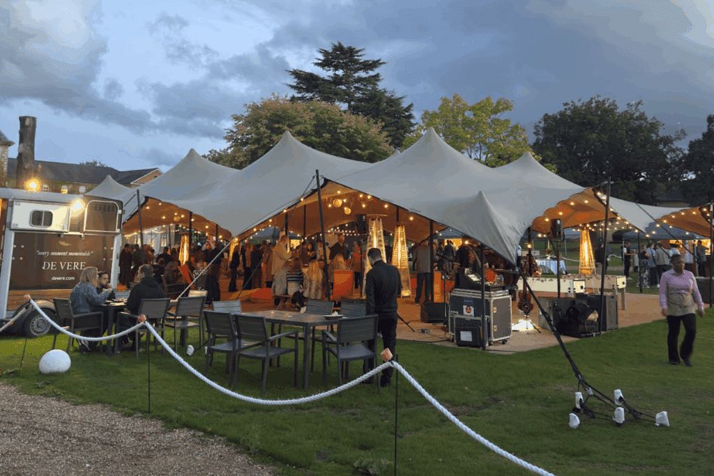 An outdoor event at dusk showcases thoughtful event strategy, with people gathered under large, illuminated canopy tents. Some guests sit at tables on the grass, while others mingle near food stalls and lights. Trees and a trailer frame the vibrant scene.