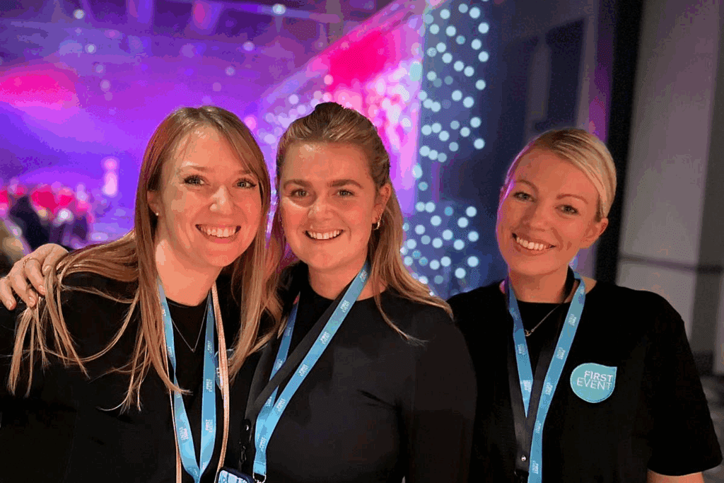 Three women wearing black outfits and light blue lanyards smile together at an indoor event careers gathering, with colorful, blurred lights in the background.