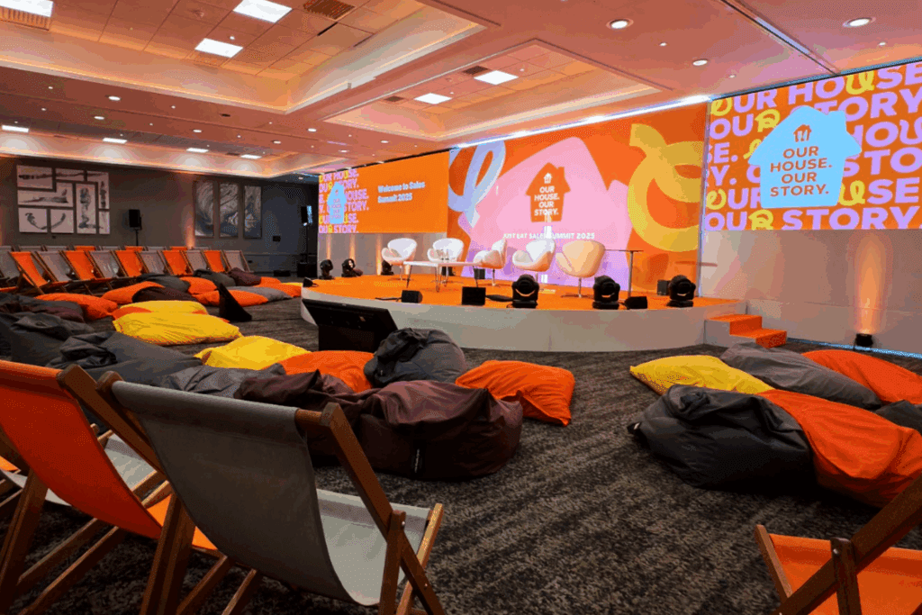 A private luxury event space featuring a stage with three chairs, colorful bean bags, and deck chairs for seating. Bright orange and yellow decor sets the scene, with OUR HOUSE. OUR STORY. displayed on screens and walls.