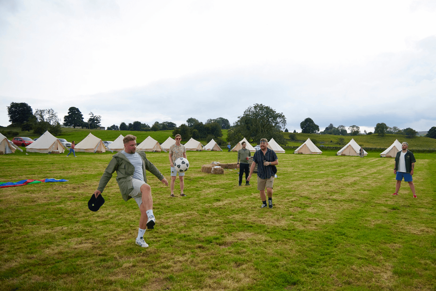 A group of people play with a large soccer ball on a grassy field with rows of tents and hay bales in the background under a cloudy sky.