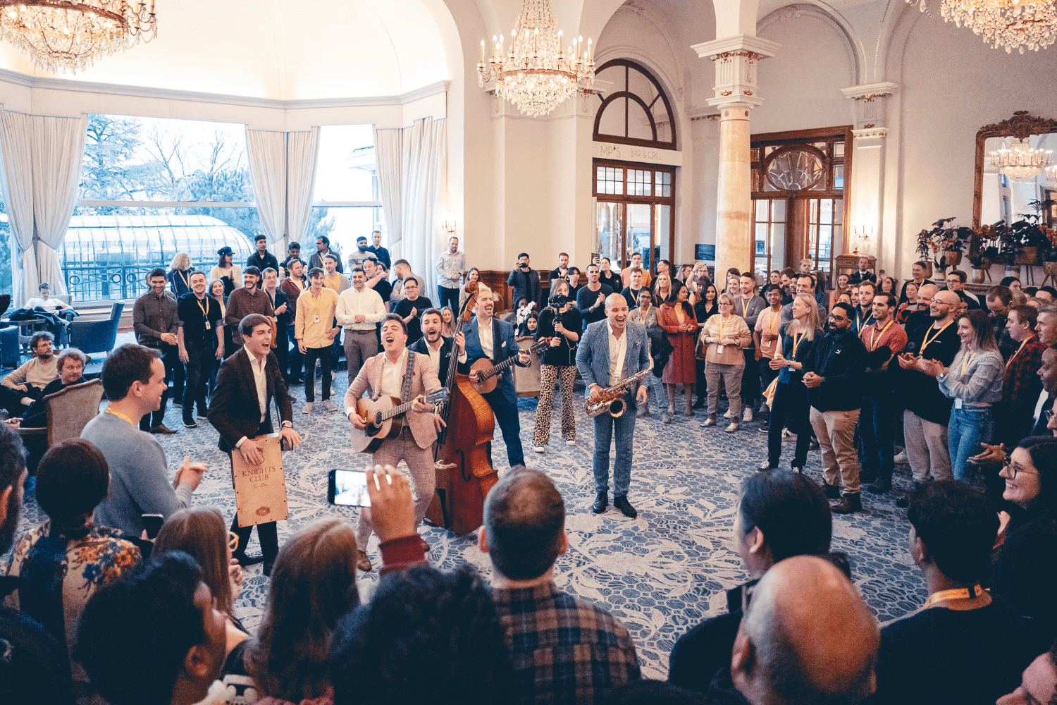 A lively band performs with guitars, a saxophone, and percussion in the center of a large, elegant room, surrounded by an enthusiastic crowd. Chandeliers hang above, and natural light streams through tall windows.