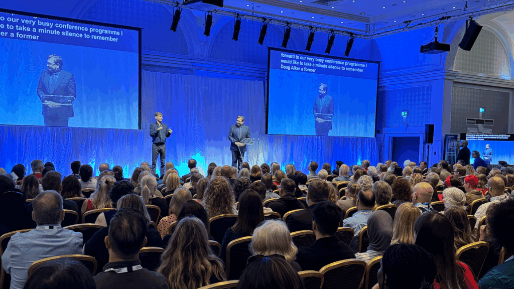 A large audience sits in a brightly lit conference hall at a Corporate Anniversary Event, facing two speakers on stage—one using sign language. A screen displays text and mirrors the scene, while decorative walls and ceiling add elegance to the occasion.