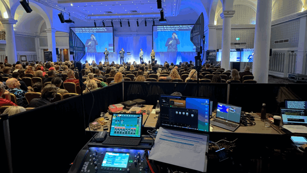 A large audience sits in an ornate hall facing a stage where several people are presenting at a Corporate Anniversary Event. In the foreground, sound and video equipment with screens and control panels are visible in the technical booth.