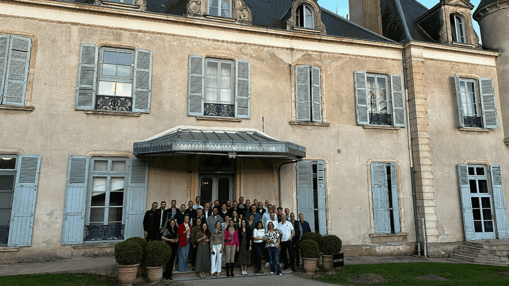 A large group of people stands in front of a historic building with light blue shutters and tall windows, posing for a group photo near the entrance under a glass awning during a sustainable corporate event.