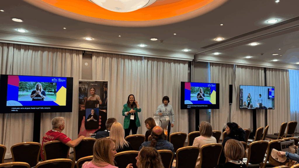 A woman and a man stand at the front of a conference room, speaking to an audience during a Corporate Anniversary Event. Two monitors display a presentation with subtitles and a woman using sign language. Rows of seated attendees face the presenters.