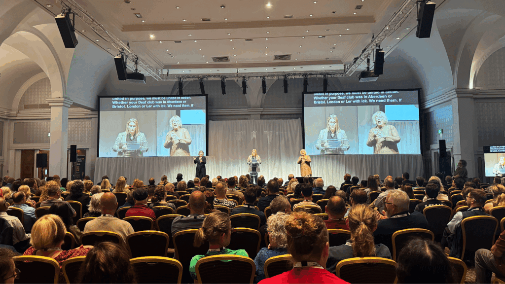A large audience sits in a conference hall at a Corporate Anniversary Event, facing a stage where three presenters stand, including one using sign language. A large screen displays captions of their speech. The room has high arched ceilings and bright lighting.