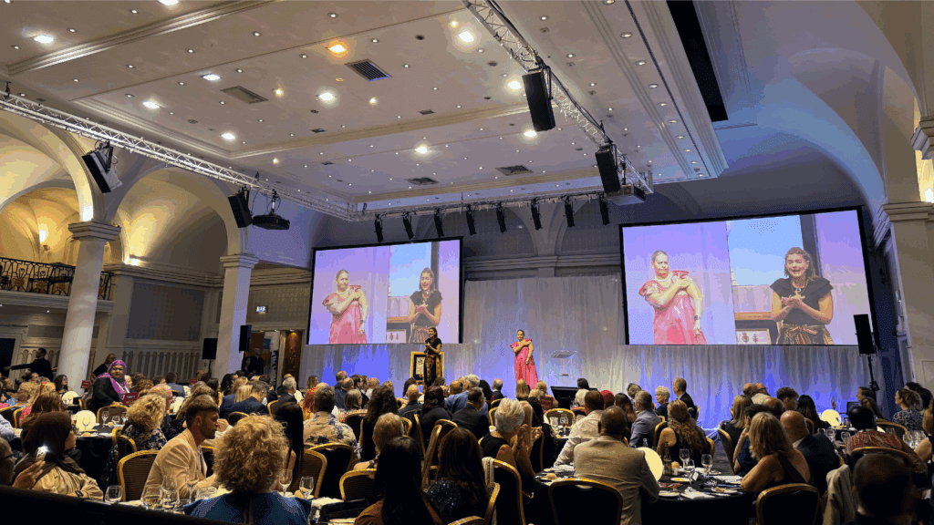 At a Corporate Anniversary Event, a woman speaks on stage at a formal gala dinner, with two large screens behind her displaying a sign language interpreter. The audience, dressed in evening attire, sits at round tables under an arched ceiling.