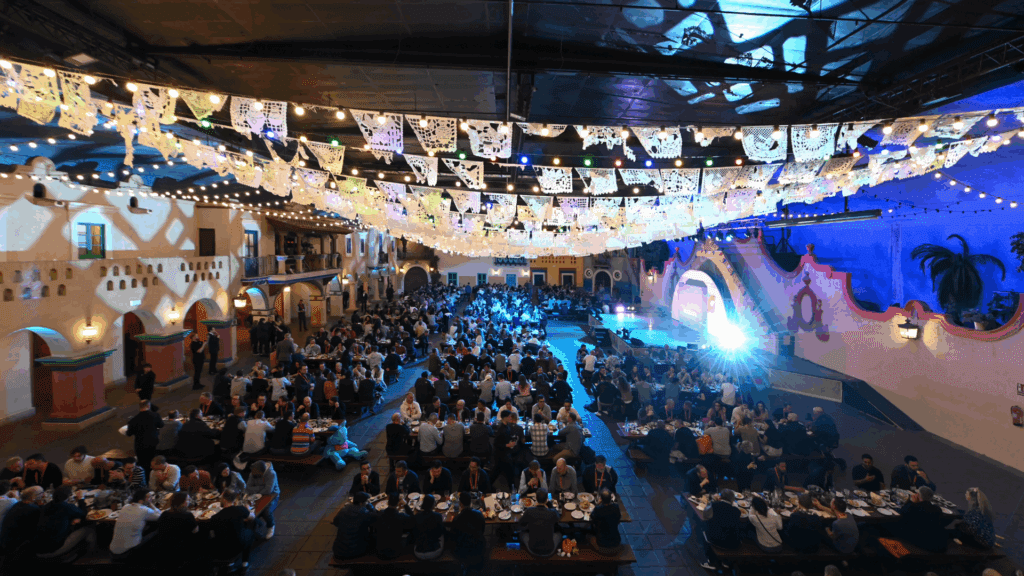 A large indoor event organised by a skilled conference organiser, with many people seated at long banquet tables, festive paper flags hanging above, and a stage lit with blue lights. The lively atmosphere feels both celebratory and professional.