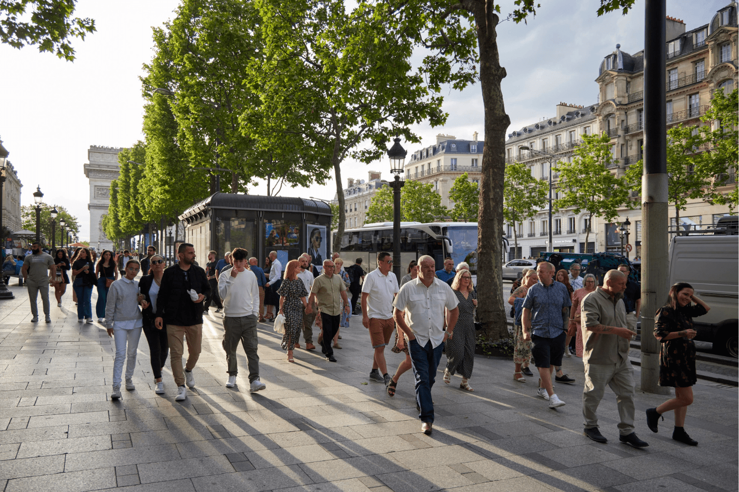 Restorative Experiences Blog. People walk along a sunny, tree-lined street in Paris near the Arc de Triomphe, enjoying restorative experiences amid lively buildings, vehicles, and a newsstand in the bustling scene.