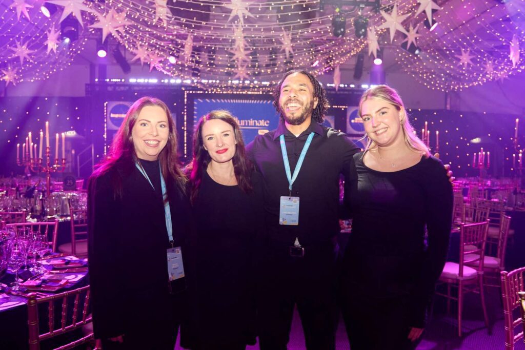 Four members of the First Event team, wearing event lanyards, stand together and smile in a decorated venue with star-shaped lights, string lights, and elegantly set tables in the background.