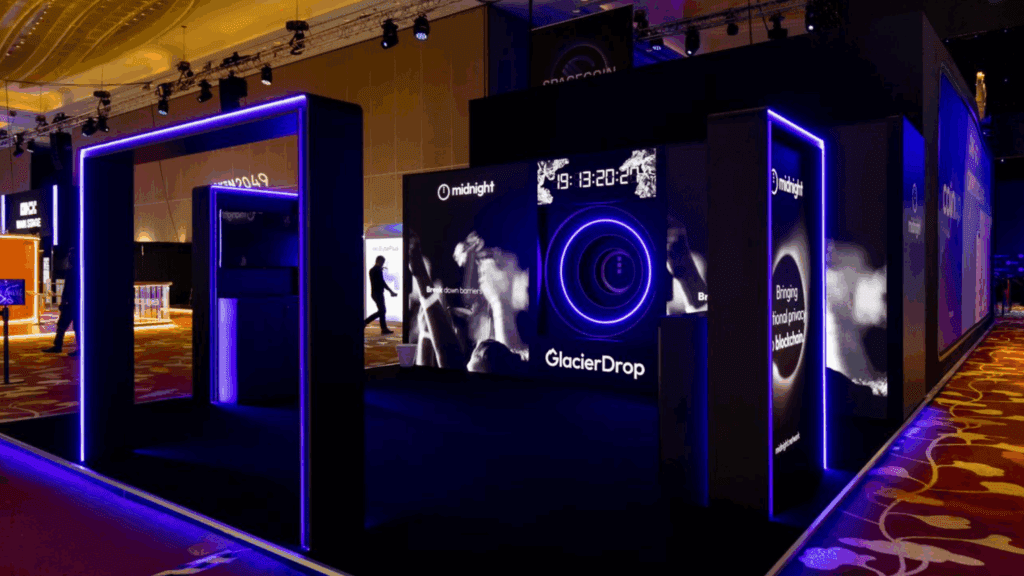 A modern exhibition booth with blue neon lights, digital displays, and the words “GlacierDrop” and “midnight,” stands out. Silhouettes of people and abstract graphics appear in the dimly lit event hall.
