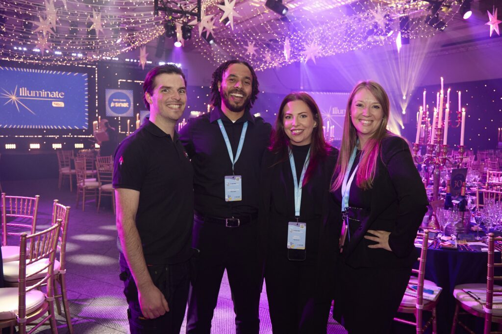 Four members of the First Event team, wearing black and event badges, stand smiling together in a decorated, star-lit venue with round tables and chairs. A large “Illuminate” sign is visible on a screen in the background.