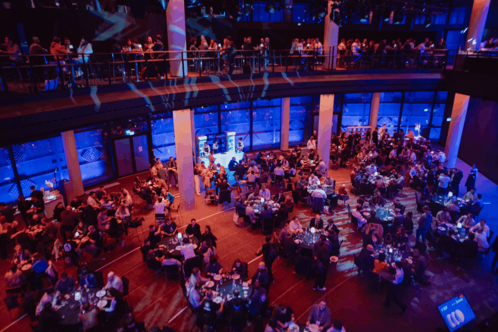 A large banquet hall filled with people seated at round tables, dining and socializing under blue lighting during the Cardano Summit, with additional guests on an upper balcony.