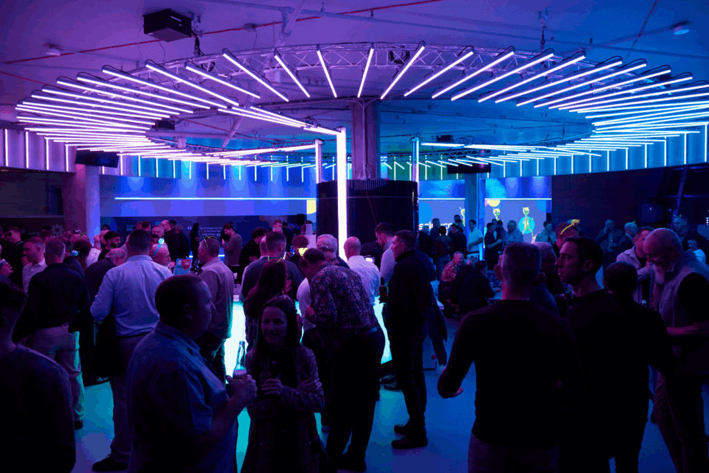 A large group of people socialize at a lively indoor event under bright, circular purple and blue neon lights, creating a vibrant atmosphere around an impressive expo stand build. Guests mingle with drinks as clusters of people converse throughout the room.