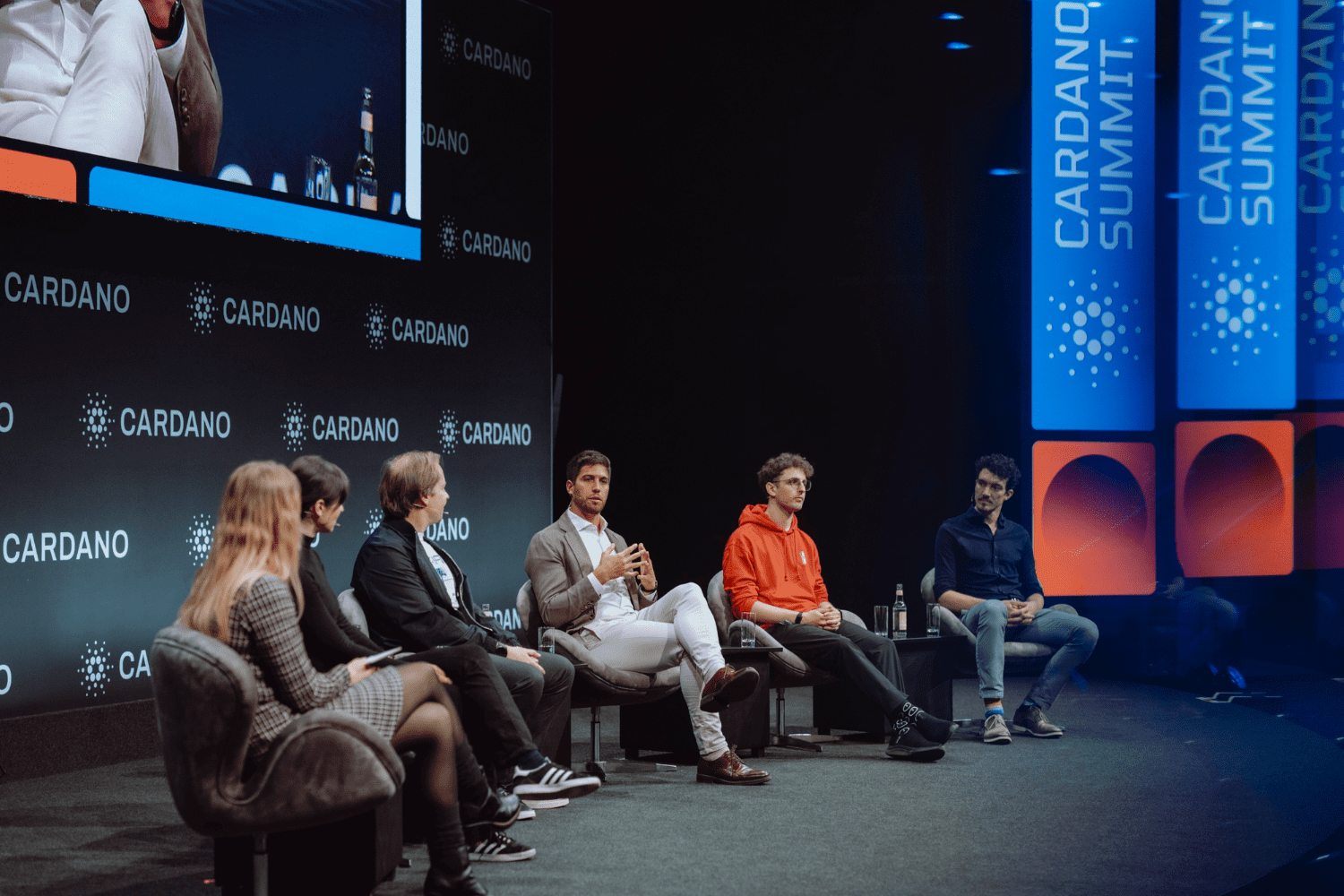 Five people sit on a stage having a panel discussion at the Cardano Summit. They face the audience, with prominent cardano summit logos displayed on the backdrop and screens behind them.