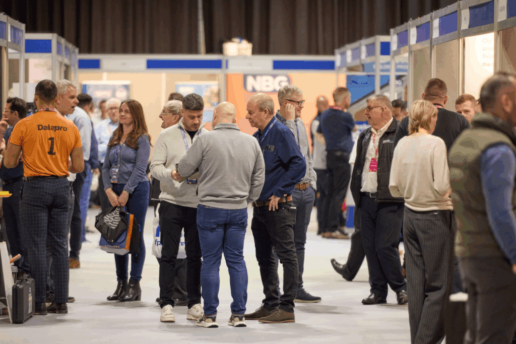 A busy indoor trade show or conference with groups of people standing, talking, and walking among expo stand build displays. Most attendees wear name badges and casual to business-casual clothing.
