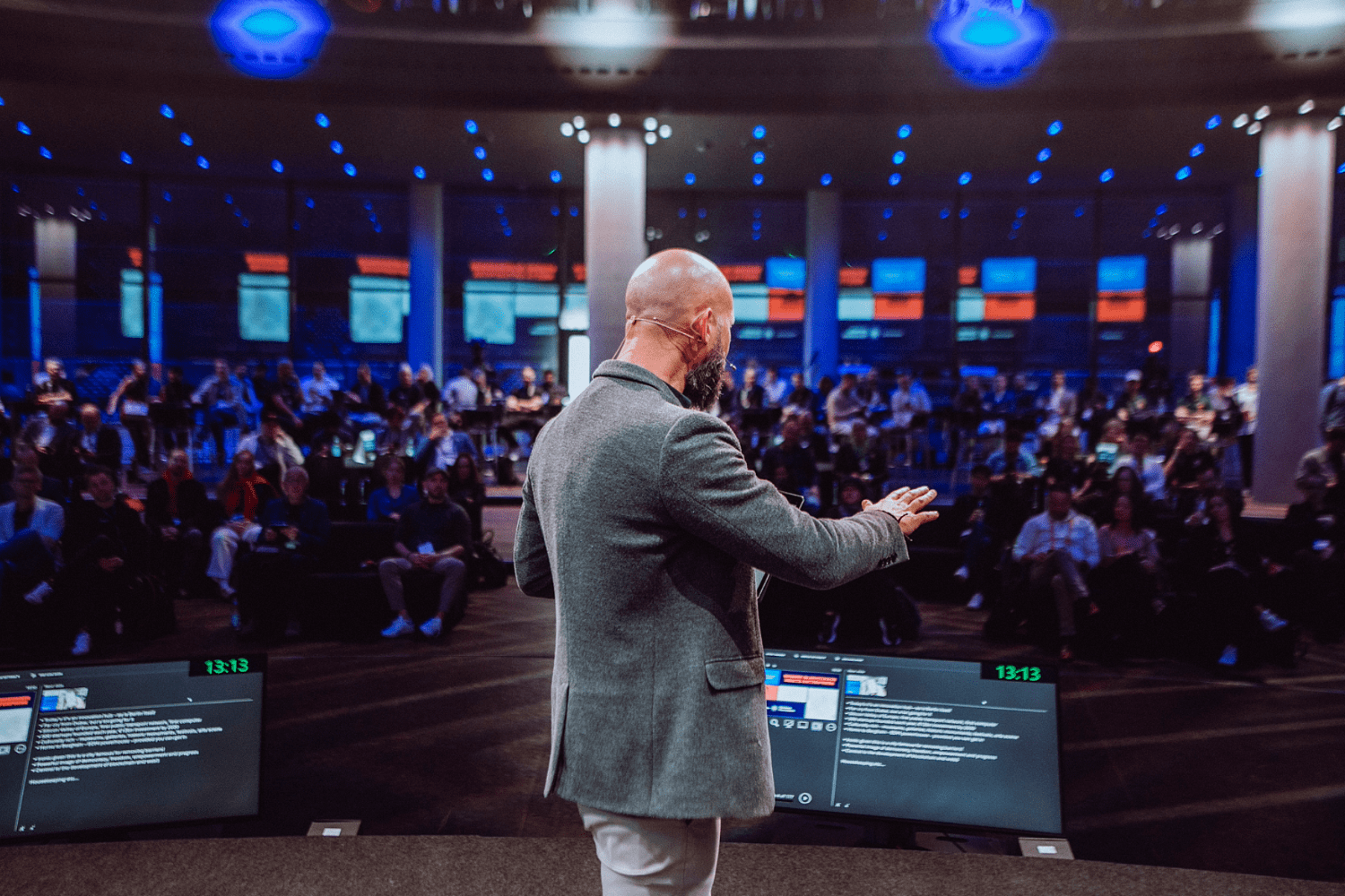 A man with a shaved head and beard, wearing a gray blazer, speaks on stage at the Cardano Summit to an audience in a large, modern conference hall with bright lights and multiple screens displaying information.