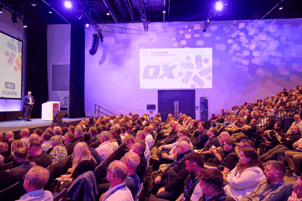 A large audience sits in a brightly lit auditorium, attentively watching a speaker on stage. Near the presentation slides, an impressive expo stand build draws interest at the front and side of the room.