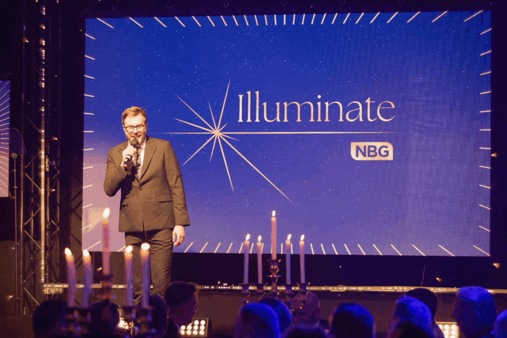 A man in a suit speaks into a microphone on stage, with a large screen displaying "Illuminate," the NBG logo, and an impressive Expo Stand Build. Candlelit tables and an attentive audience are visible in the foreground.