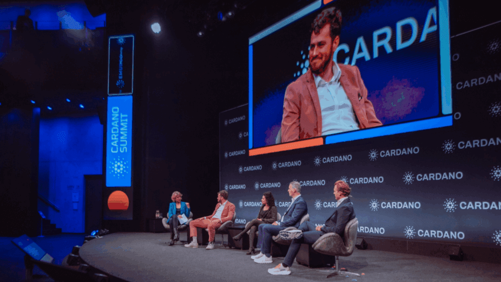 A panel of five people sits on stage at the Cardano Summit, with a large screen behind them showing one panelist speaking. The stage and backdrop prominently feature the Cardano logo.
