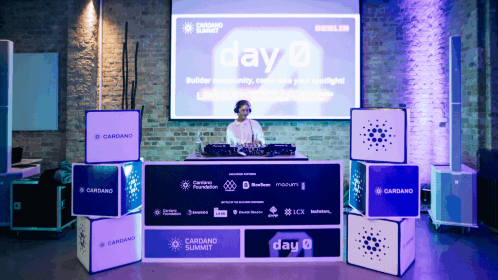 A DJ stands behind a mixing console at an event with glowing Cardano Summit signs, cube displays, and a projected Day 0 Cardano Summit presentation on a brick wall in a modern, vibrant venue.