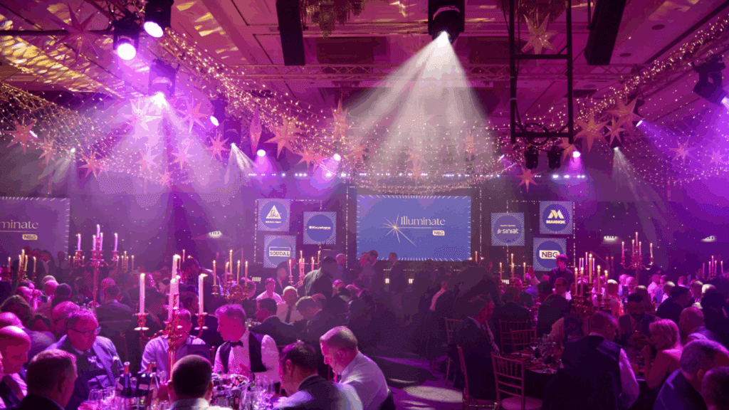 A large, elegant banquet hall filled with people at round tables, lit by purple stage lights and candle centerpieces. A stage featuring an Expo Stand Build and a screen reading 