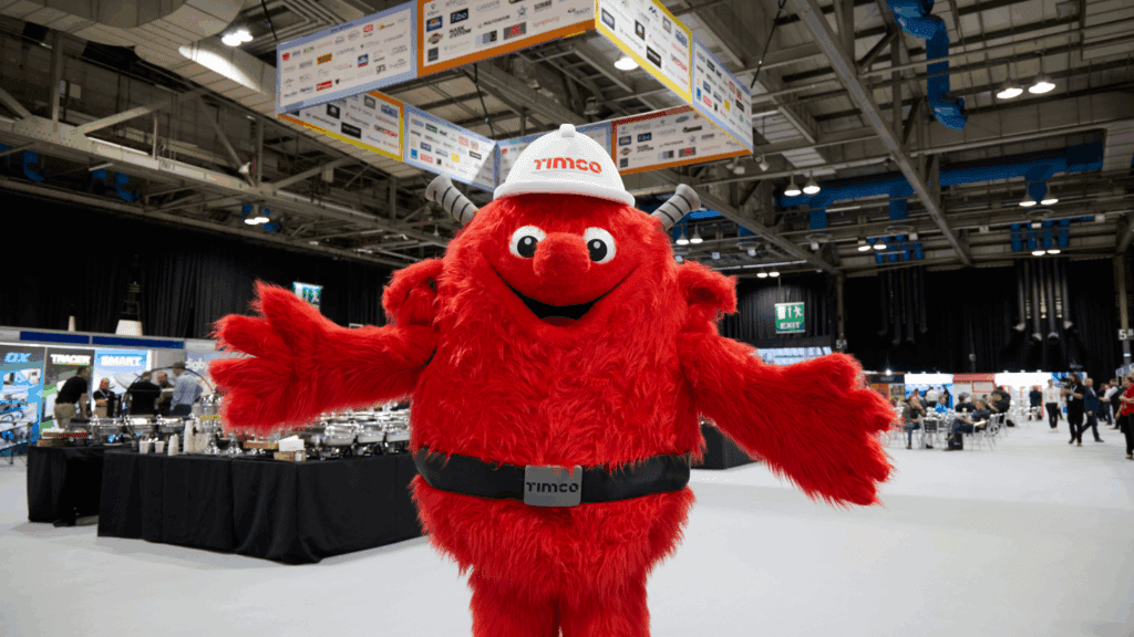 A person in a large, red, furry monster costume with a TIMCO hard hat stands with arms open at an indoor expo stand build or trade show. Exhibition booths and people are visible in the background.
