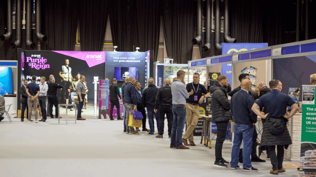 People interact at various booths during an indoor trade show or exhibition, featuring custom Expo Stand Build designs. Some are standing and conversing, while others browse displays amid promotional banners and informational signs.