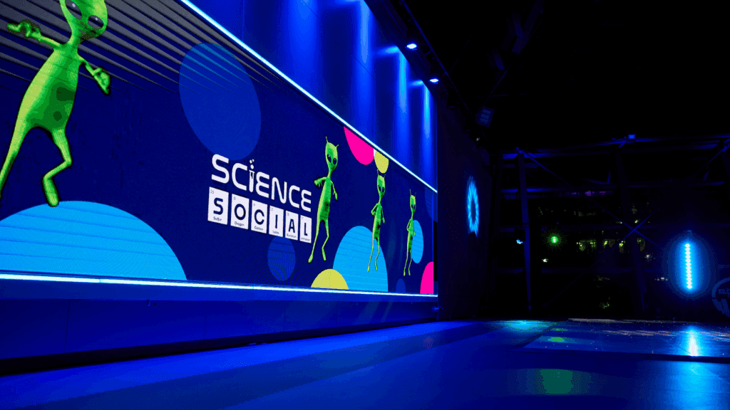 A large digital screen at the Expo Stand Build displays green cartoon aliens and the words SCIENCE SOCIAL in a futuristic, blue-lit room filled with colorful shapes in the background.