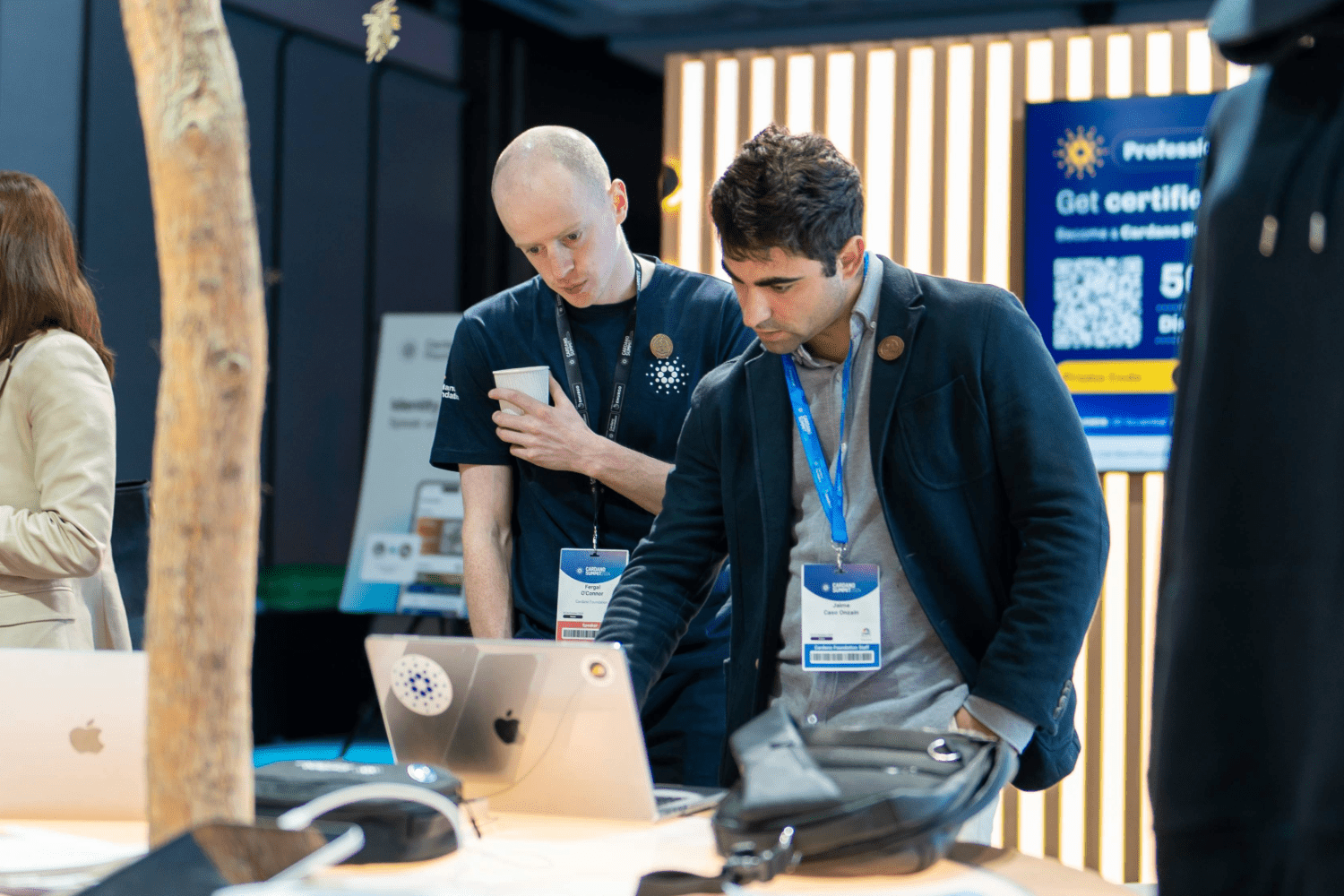 Two men at a tech event stand at a table with laptops. One checks his phone as the other points at a laptop screen featuring webinars on-demand. Both wear event badges and appear focused. A promotional sign is visible in the background.