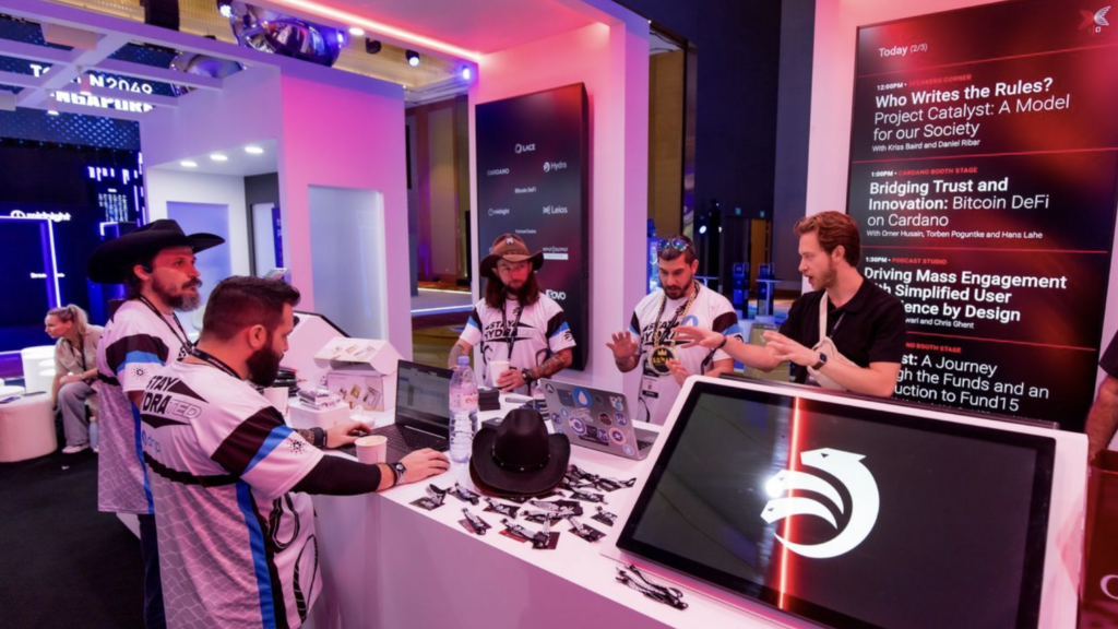 Five men in matching white shirts gather around an expo stand build at a tech conference. Branded swag, laptops, and a cowboy hat are on the table as digital screens display event schedules and a stylized bird logo in the background.