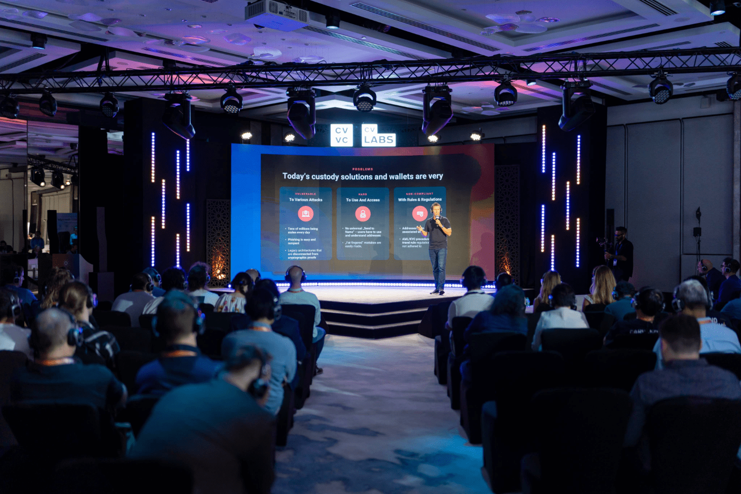 A speaker stands on stage presenting creative event services to an attentive audience in a conference room, with a large screen behind displaying information about custody solutions and wallets.