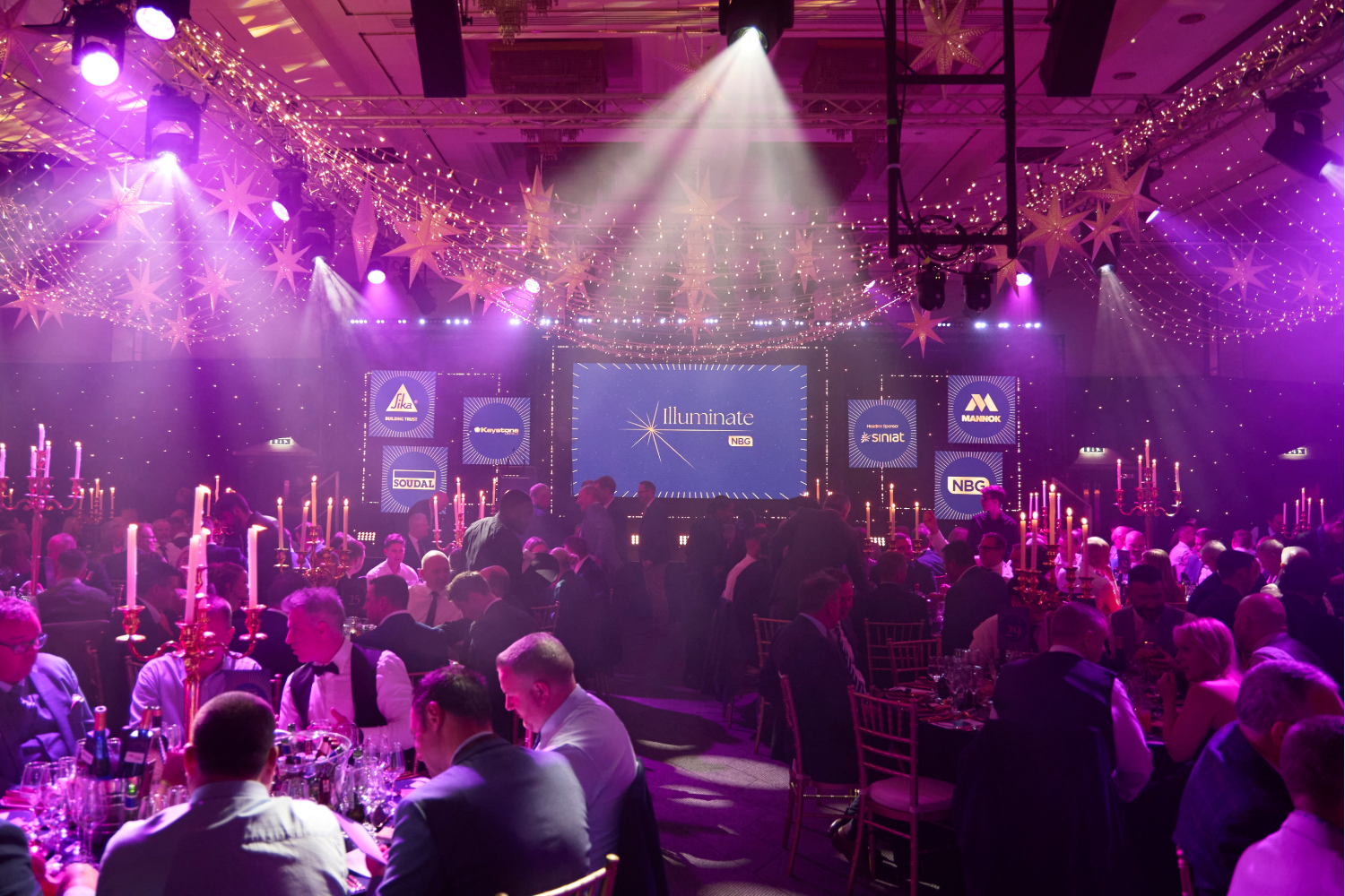 A large crowd sits at round tables with tall candle centerpieces in a dimly lit banquet hall, illuminated by purple stage lights and spotlights, facing a stage with screens displaying logos and a striking Expo Stand Build as the focal point.