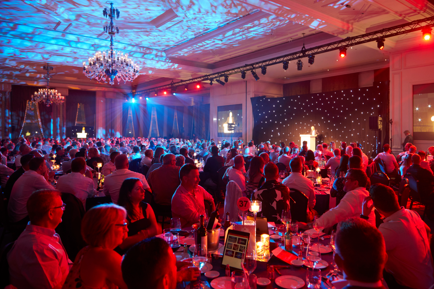 A large crowd sits at round tables in a ballroom with chandeliers, listening to a speaker at a lectern on a stage with starry lights. The room is lit with red and blue ambient lighting.