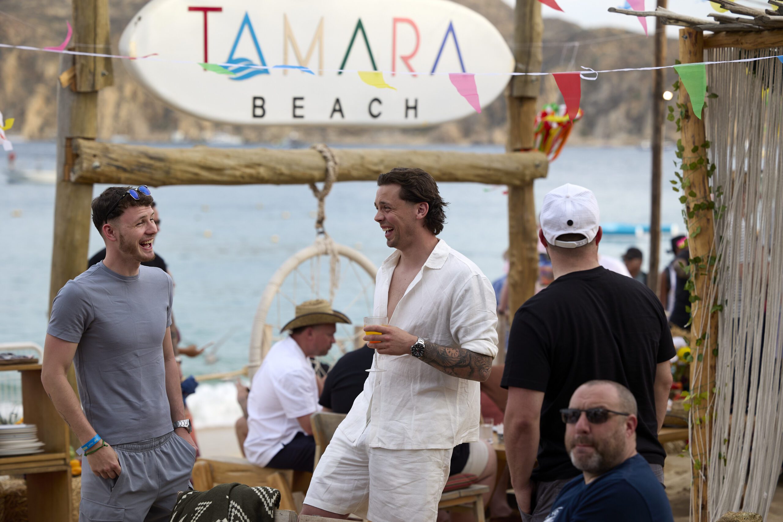 Several men socialise and smile at a lively beachside venue with a “Tamara Beach” sign overhead. The scene, reminiscent of an industry conference after-party, features colourful decorations, people relaxing, and the sea in the background.